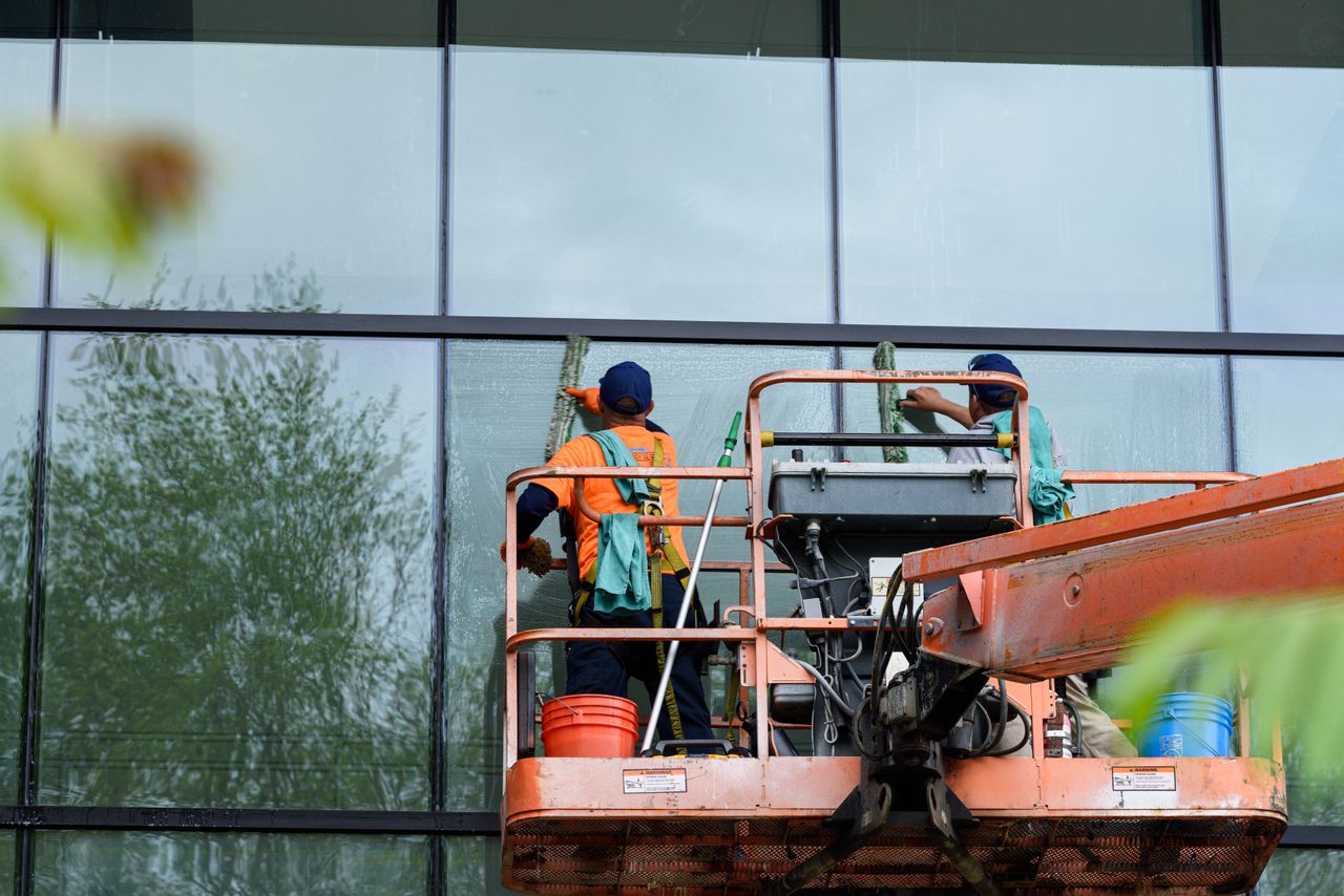 Two workers wash windows on a glass building facade using a lift platform, improving the building's appearance.