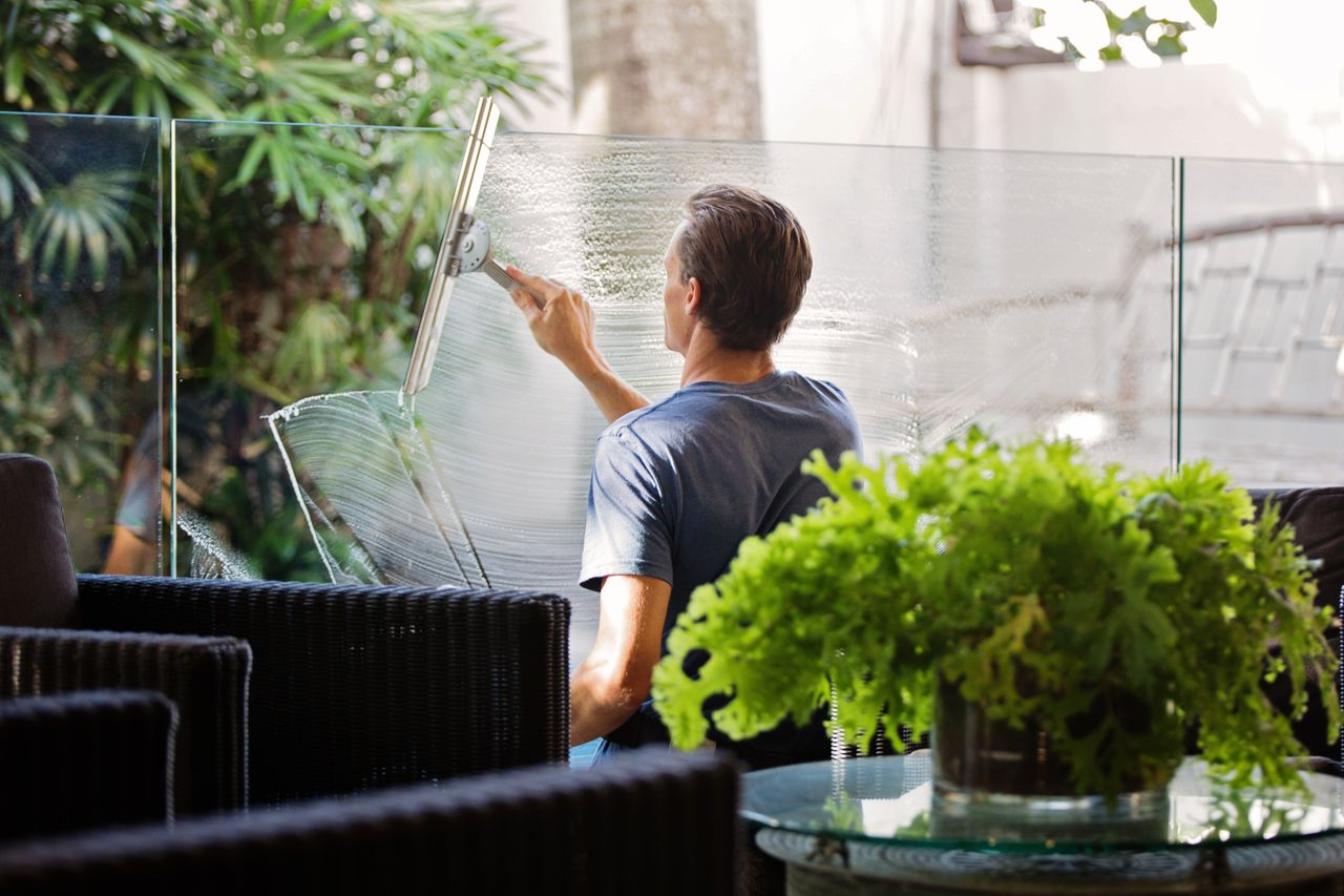 A man outdoors cleaning a glass barrier with a squeegee, with greenery all around.