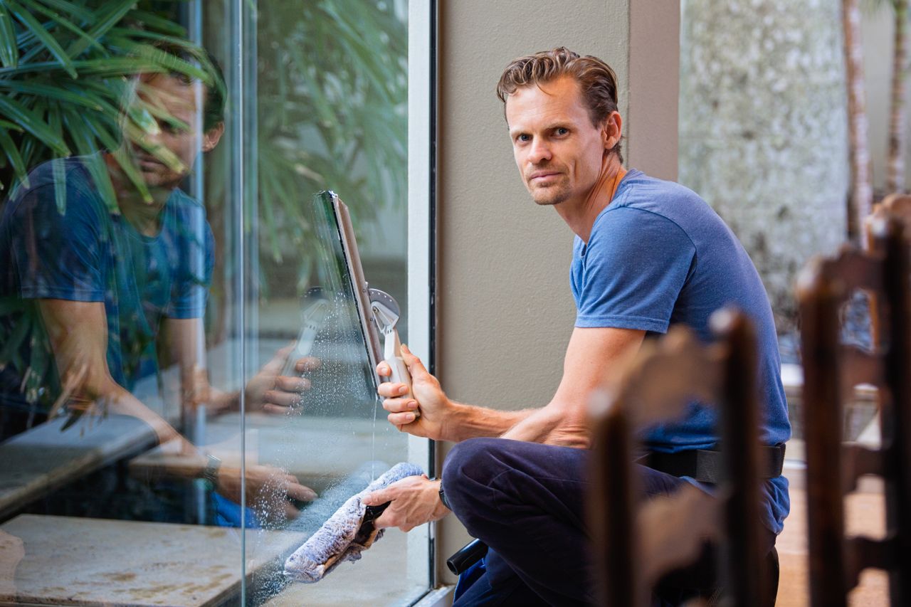 why-choose-us A man in a blue t-shirt cleaning large glass pane with a squeegee indoors.